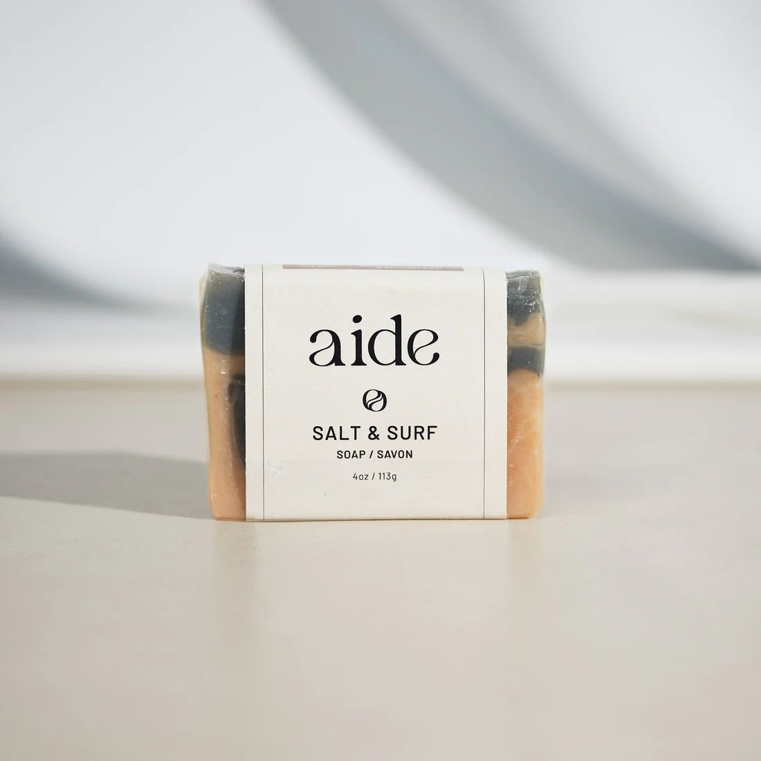 Upright light pink and grey bar of soap with a white band label with black text and black logos, with gold detailing