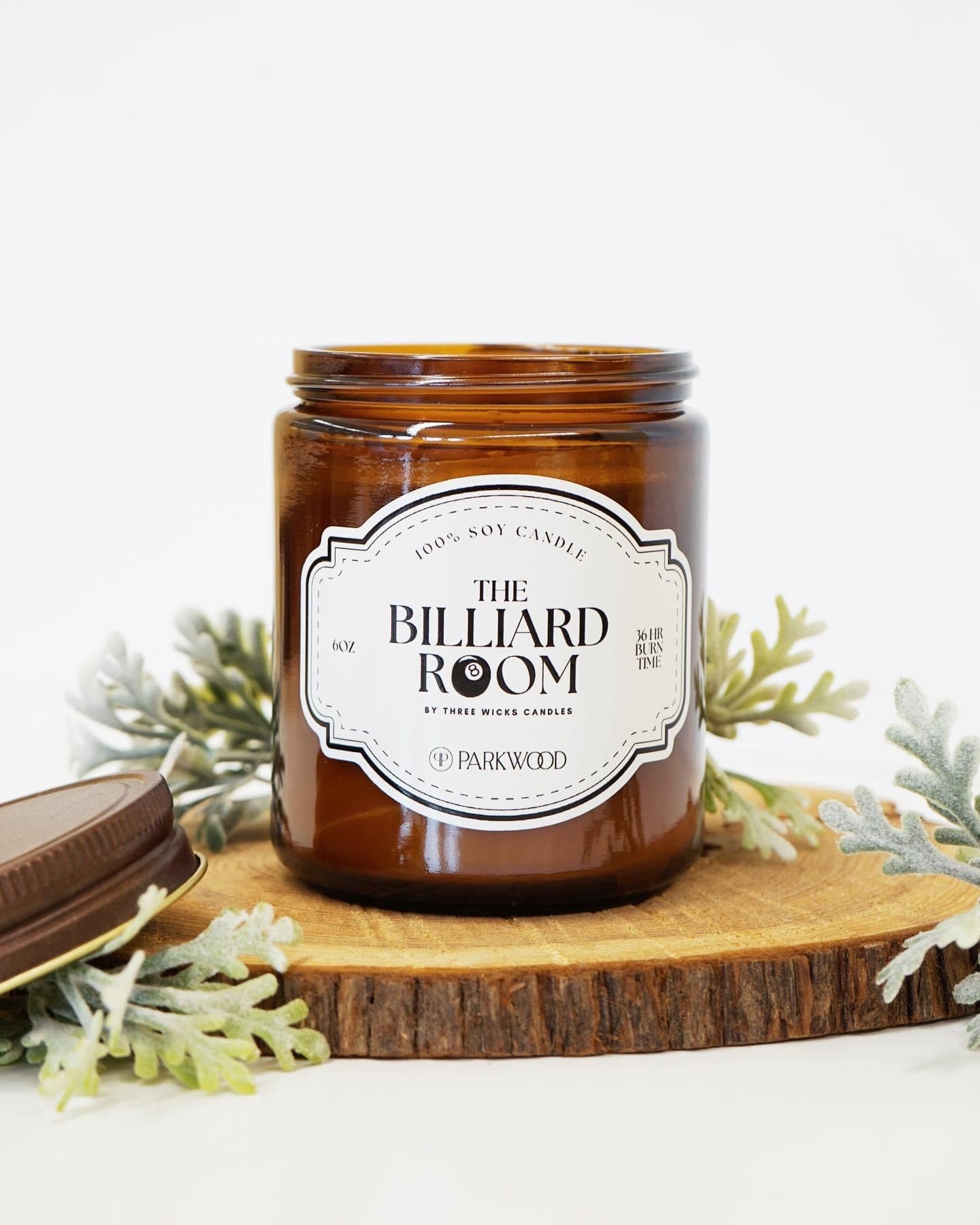 Brown glass candle jar with a label that says "The Billiard Room" with an 8 ball as one of the o's surrounded by wood, and herb leaves