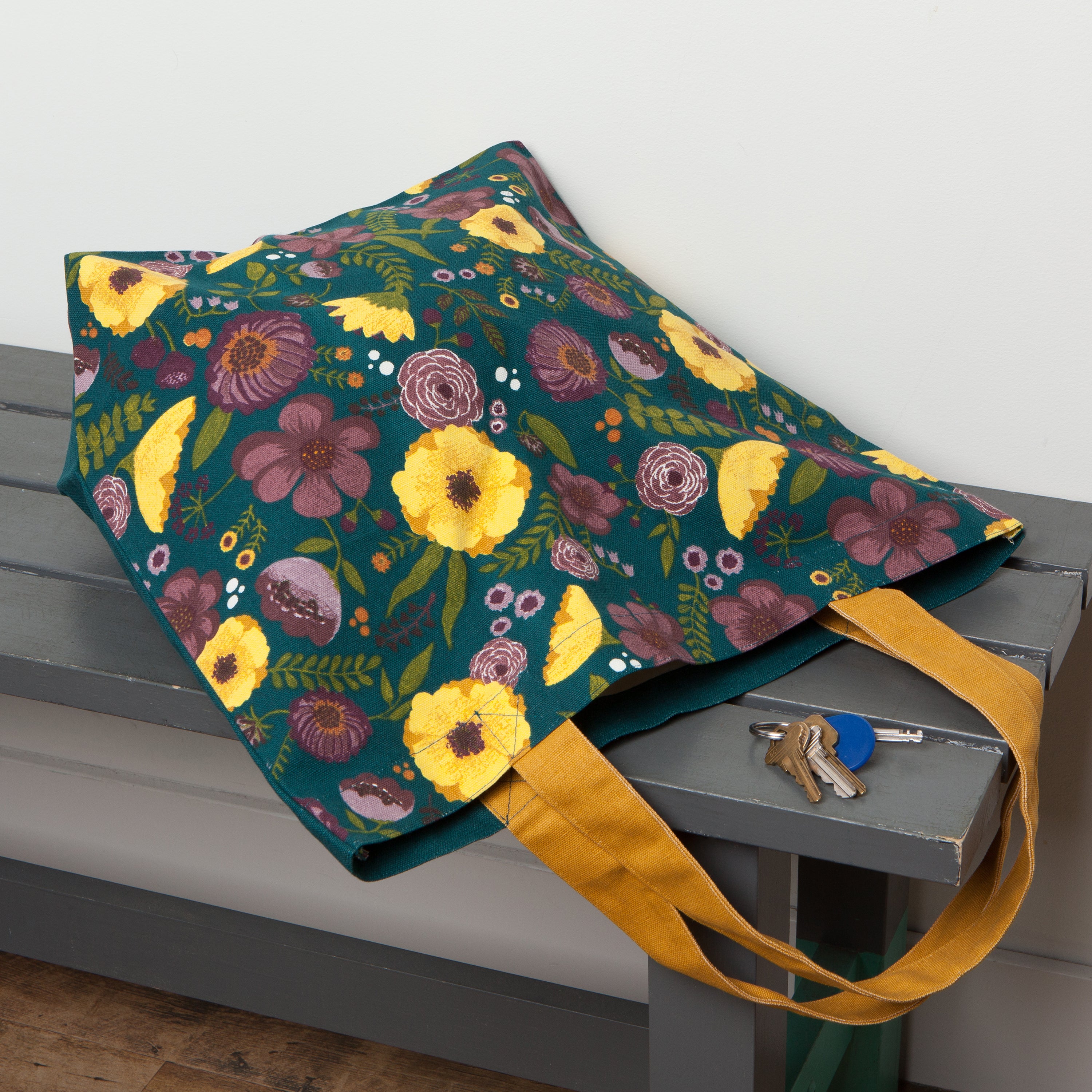 dark green tote bag with yellow handles, yellow and purple flowers with green stems on a grey bench with keys next to it