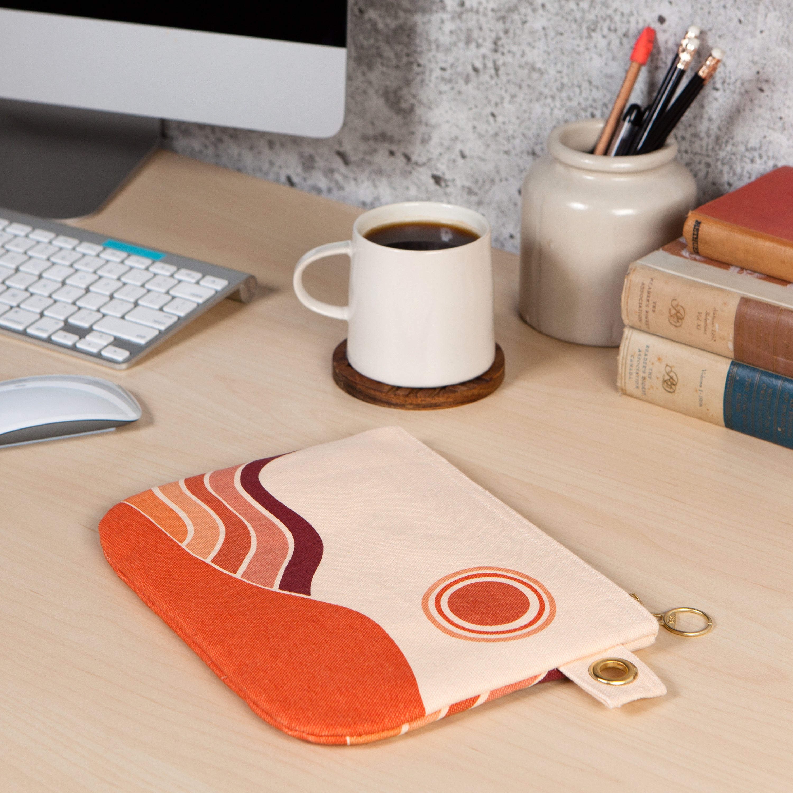 an Off white zip up pouch with a orange palleted simplistic beach landscape  next to a home computer and cup of coffee