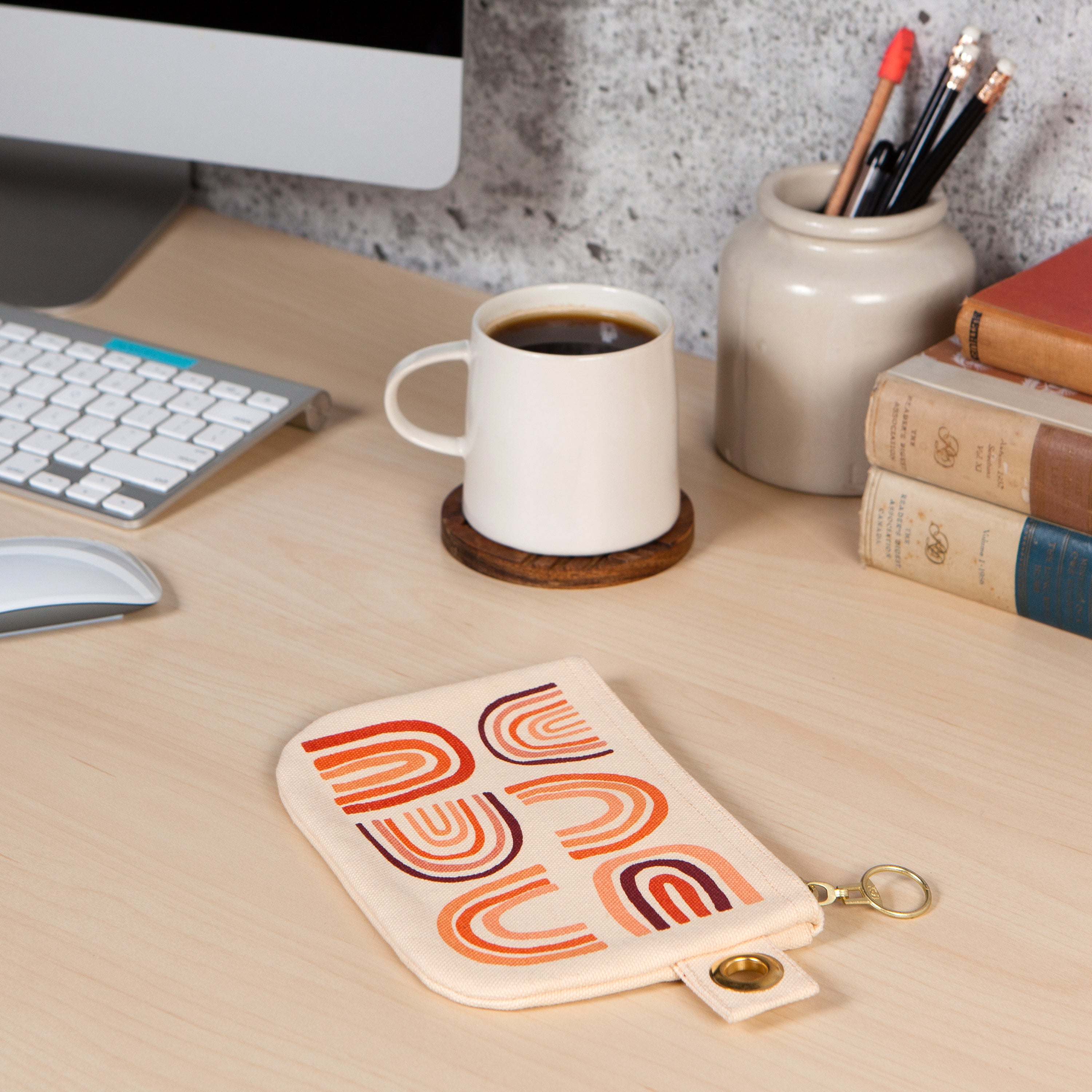 Beige pouch with gold zipper and a rainbow of various shades of orange on a computer desk with a cup of coffee next to it