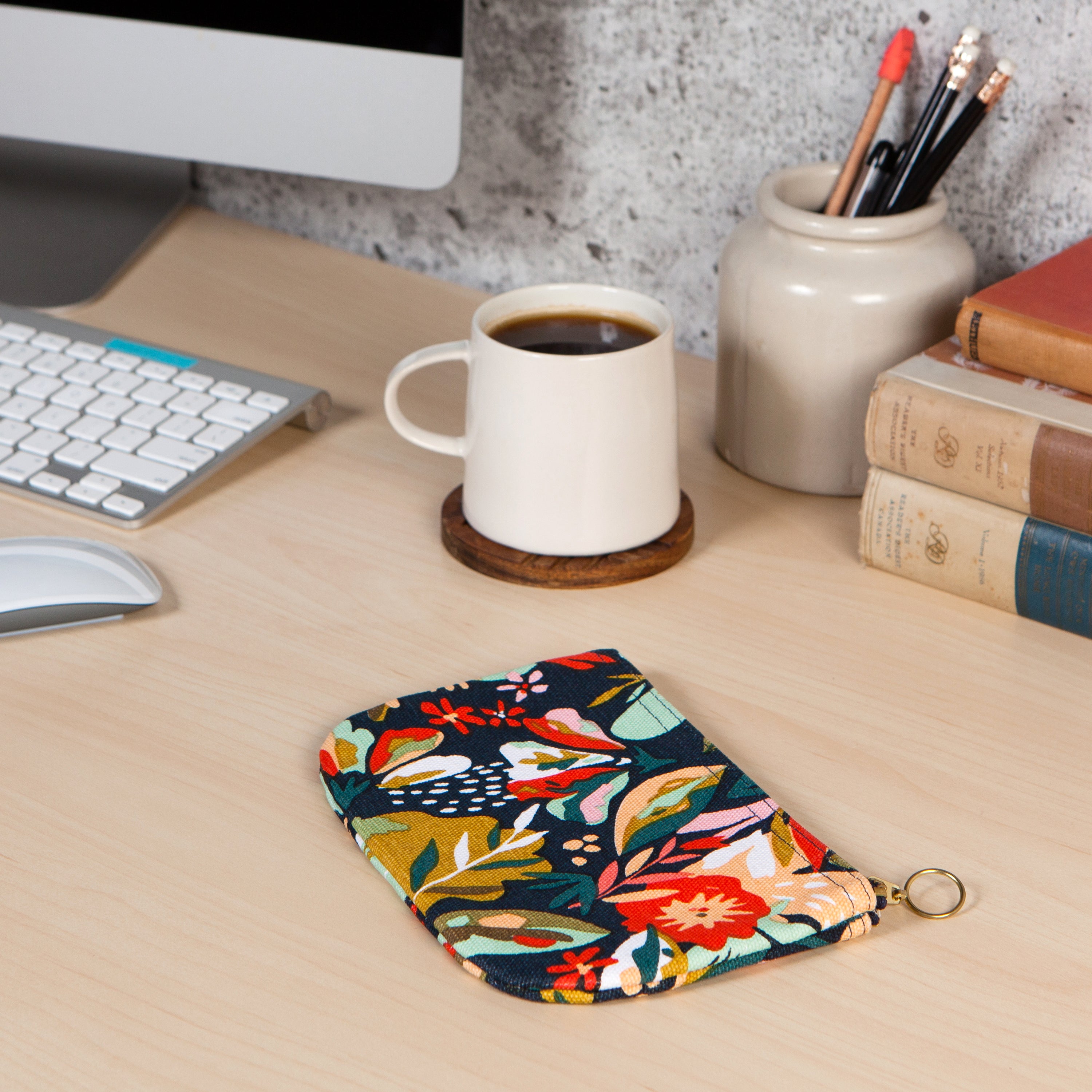 Blue pouch with gold zipper and multi coloured flower pattern on a computer desk with a cup of coffee next to It