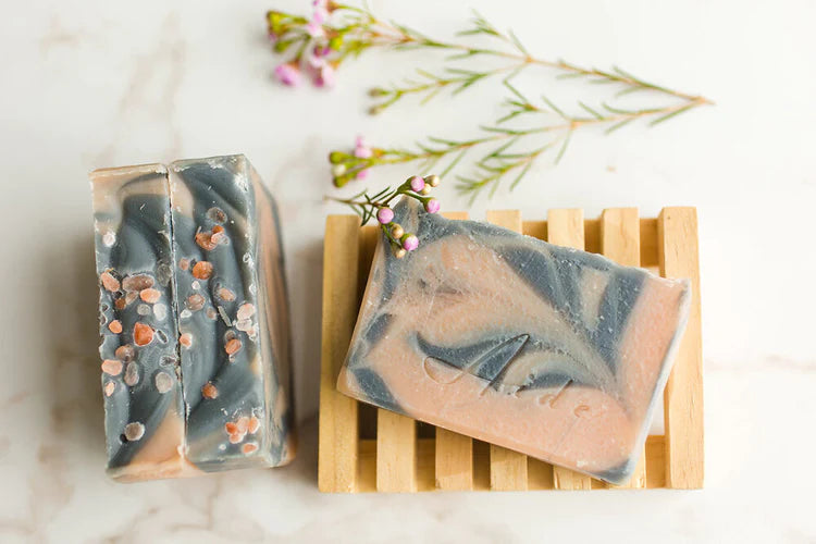 Three grey and pink bars of soap, with some flowers and a wooden rack surrounding them