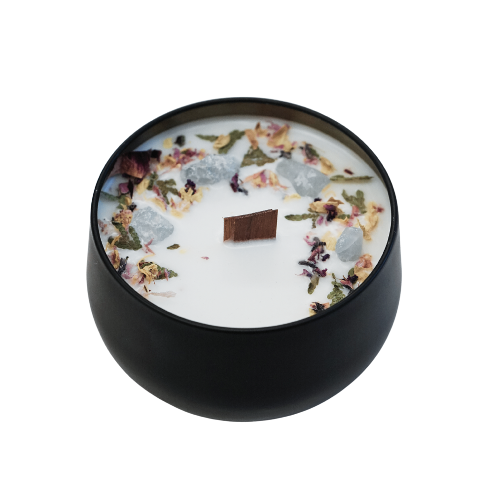 white candle with different herbs and leaves inside a short black metal candle jar