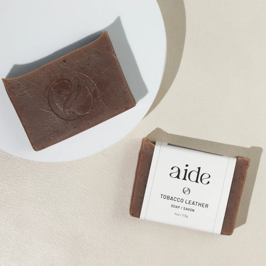 Two bars of reddish-brown soap, one on a white plate, while the other has a white band label