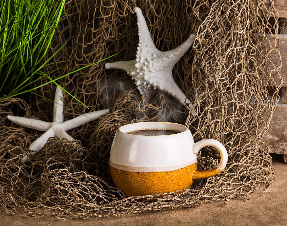 White and brown round textured mug next to a fishing net, starfish, and sea grass