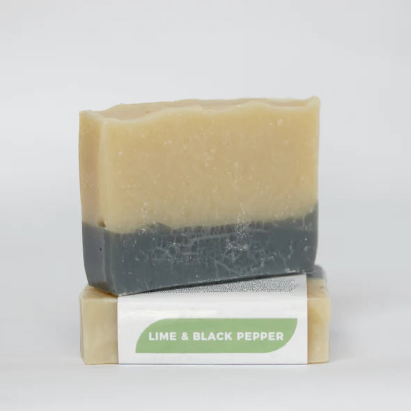 Two black and beige bars of soap, one is upright and the other has a white band label with a green logo that says "lime and black pepper"