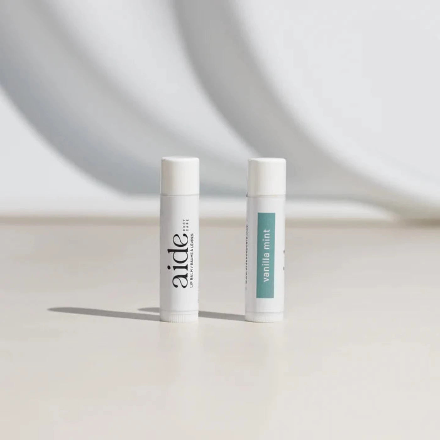 Two side by side white lip balm containers, with one showing the Aide wordmark, and the other showing a teal rectangle that reads "Vanilla Mint"
